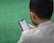 A man is sitting on a green surface, reading content on a smartphone. The text on the phone's screen is in Arabic script.