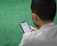 A man is sitting on a green surface, reading content on a smartphone. The text on the phone's screen is in Arabic script.