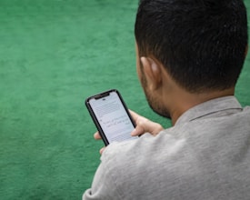 A man is sitting on a green surface, reading content on a smartphone. The text on the phone's screen is in Arabic script.