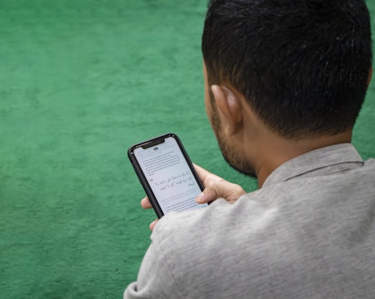 A man is sitting on a green surface, reading content on a smartphone. The text on the phone's screen is in Arabic script.