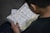 A person is reading an open book containing text in Arabic script. The page layout includes tables and possibly a commentary or translation in a smaller font at the bottom. The person is seated and likely engaged in studying or reading.