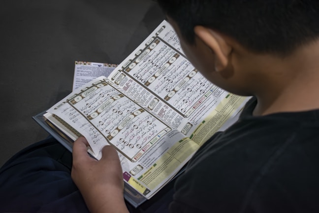 A person is reading an open book containing text in Arabic script. The page layout includes tables and possibly a commentary or translation in a smaller font at the bottom. The person is seated and likely engaged in studying or reading.