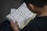 A person is reading an open book containing text in Arabic script. The page layout includes tables and possibly a commentary or translation in a smaller font at the bottom. The person is seated and likely engaged in studying or reading.