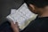 A person is reading an open book containing text in Arabic script. The page layout includes tables and possibly a commentary or translation in a smaller font at the bottom. The person is seated and likely engaged in studying or reading.