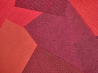 A collage of fabric pieces in varying shades of red, arranged in overlapping geometric patterns. Textures of the fabrics are visible, contributing to a layered and tactile appearance.