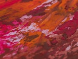 Abstract painting featuring vibrant colors and textured brushstrokes. The dominant hues include shades of red, orange, and pink intermingling with touches of white and brown. The composition emphasizes a rich, dynamic energy through the use of bold streaks and patches of color.