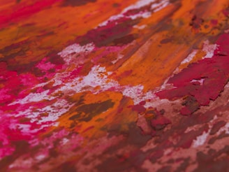 Abstract painting featuring vibrant colors and textured brushstrokes. The dominant hues include shades of red, orange, and pink intermingling with touches of white and brown. The composition emphasizes a rich, dynamic energy through the use of bold streaks and patches of color.