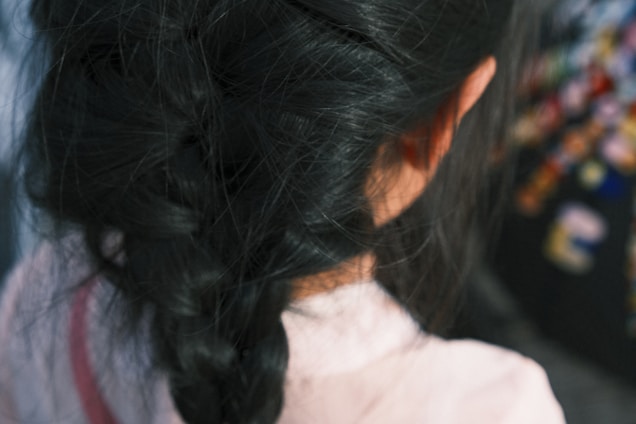 A close-up view of a person with dark braided hair from behind. The focus is on the intricately woven braid, with a blurred background featuring colorful, indistinct shapes.