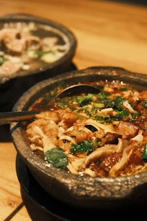 Two stone bowls filled with savory soup, garnished with various ingredients including fresh herbs and sliced vegetables. One bowl showcases a rich, reddish broth, while the other has a lighter, clearer soup with chunks of meat.