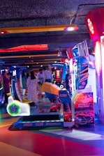 An arcade setting with a variety of brightly colored racing game machines. Several people are seen interacting in the background, and the environment is illuminated with neon lights in shades of blue, red, and purple.