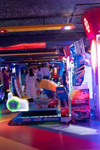 An arcade setting with a variety of brightly colored racing game machines. Several people are seen interacting in the background, and the environment is illuminated with neon lights in shades of blue, red, and purple.