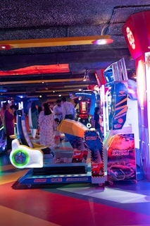 An arcade setting with a variety of brightly colored racing game machines. Several people are seen interacting in the background, and the environment is illuminated with neon lights in shades of blue, red, and purple.