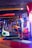 An arcade setting with a variety of brightly colored racing game machines. Several people are seen interacting in the background, and the environment is illuminated with neon lights in shades of blue, red, and purple.