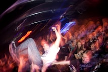 A lively, crowded concert scene with a person being lifted above the audience, suggesting crowd surfing. The lighting is dynamic with a mix of vibrant colors, creating a sense of motion and energy throughout the crowd.