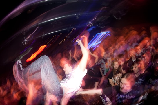 A lively, crowded concert scene with a person being lifted above the audience, suggesting crowd surfing. The lighting is dynamic with a mix of vibrant colors, creating a sense of motion and energy throughout the crowd.