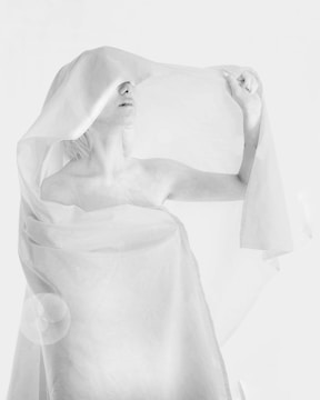 A person draped in a translucent fabric covers their eyes, creating a dreamy and ethereal atmosphere. The soft, flowing material adds texture and depth, while the monochrome palette enhances the minimalistic and artistic feel.