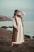 A person wearing a long, flowing white dress with a brown vest stands on a sandy beach. They appear to be looking into the distance, with one hand raised to shield their eyes. The background features a calm sea and a rocky coastline under a clear sky.
