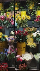 A vibrant display of various flowers arranged in yellow metal containers. The assortment includes sunflowers, red roses, lilies, and other colorful blooms. The containers are labeled 'TEX AS 1895' and are surrounded by greenery and additional flower bouquets. The setting appears to be a florist shop with an array of fresh flowers organized neatly.