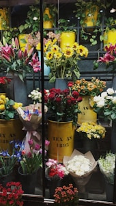 A vibrant display of various flowers arranged in yellow metal containers. The assortment includes sunflowers, red roses, lilies, and other colorful blooms. The containers are labeled 'TEX AS 1895' and are surrounded by greenery and additional flower bouquets. The setting appears to be a florist shop with an array of fresh flowers organized neatly.