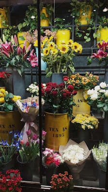 A vibrant display of various flowers arranged in yellow metal containers. The assortment includes sunflowers, red roses, lilies, and other colorful blooms. The containers are labeled 'TEX AS 1895' and are surrounded by greenery and additional flower bouquets. The setting appears to be a florist shop with an array of fresh flowers organized neatly.