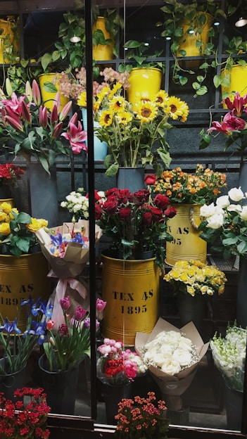 A vibrant display of various flowers arranged in yellow metal containers. The assortment includes sunflowers, red roses, lilies, and other colorful blooms. The containers are labeled 'TEX AS 1895' and are surrounded by greenery and additional flower bouquets. The setting appears to be a florist shop with an array of fresh flowers organized neatly.
