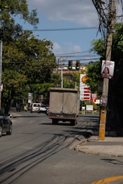 A busy urban street features vehicles including a truck and several cars moving along the road. Overhead traffic lights show a green signal, and power lines stretch across the scene. A Shell gas station with prices displayed is visible in the background, and lush green trees line the street, providing shade. A billboard attached to a pole depicts a person and signs of urban wear such as a cracked sidewalk can be observed.