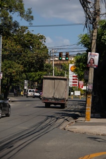 A busy urban street features vehicles including a truck and several cars moving along the road. Overhead traffic lights show a green signal, and power lines stretch across the scene. A Shell gas station with prices displayed is visible in the background, and lush green trees line the street, providing shade. A billboard attached to a pole depicts a person and signs of urban wear such as a cracked sidewalk can be observed.