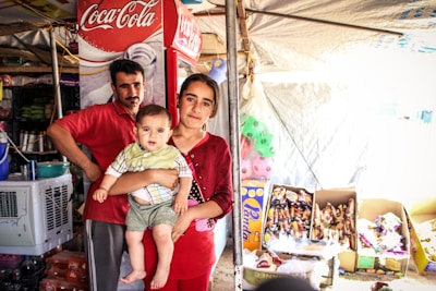 A family stands in a small shop or market stall. A man and a woman hold a baby. The stall is filled with various items, including beverages and snacks. A refrigerator with a Coca-Cola logo is visible in the background.