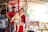 A family stands in a small shop or market stall. A man and a woman hold a baby. The stall is filled with various items, including beverages and snacks. A refrigerator with a Coca-Cola logo is visible in the background.