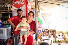 A family stands in a small shop or market stall. A man and a woman hold a baby. The stall is filled with various items, including beverages and snacks. A refrigerator with a Coca-Cola logo is visible in the background.