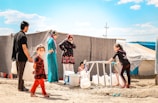 Several people are gathered around a water distribution point in what appears to be a refugee camp. A young girl and a man are carrying large white containers, while other women and children are waiting nearby. Tents and makeshift shelters can be seen in the background, suggesting a temporary living situation.