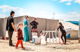 Several people are gathered around a water distribution point in what appears to be a refugee camp. A young girl and a man are carrying large white containers, while other women and children are waiting nearby. Tents and makeshift shelters can be seen in the background, suggesting a temporary living situation.