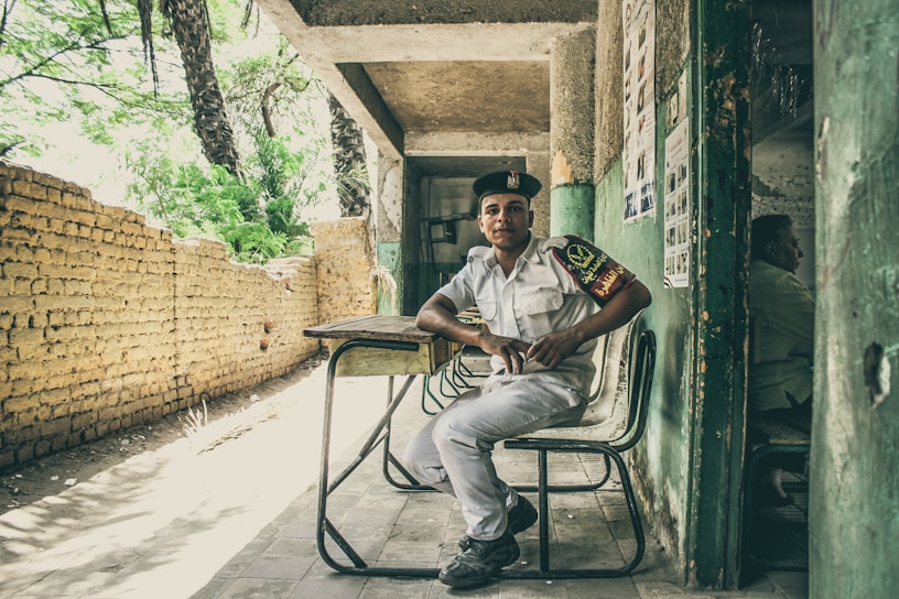 A young man in a uniform is seated on a chair in front of a makeshift desk, positioned in a covered area that appears to be partially outdoors. The background features a rustic brick wall and lush green trees, while posters are visible on the wall beside him. The atmosphere feels casual and relaxed.