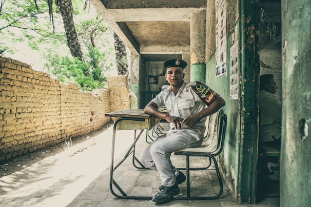 A young man in a uniform is seated on a chair in front of a makeshift desk, positioned in a covered area that appears to be partially outdoors. The background features a rustic brick wall and lush green trees, while posters are visible on the wall beside him. The atmosphere feels casual and relaxed.