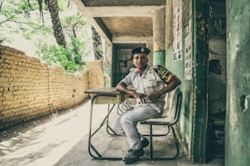 A young man in a uniform is seated on a chair in front of a makeshift desk, positioned in a covered area that appears to be partially outdoors. The background features a rustic brick wall and lush green trees, while posters are visible on the wall beside him. The atmosphere feels casual and relaxed.