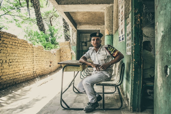 A young man in a uniform is seated on a chair in front of a makeshift desk, positioned in a covered area that appears to be partially outdoors. The background features a rustic brick wall and lush green trees, while posters are visible on the wall beside him. The atmosphere feels casual and relaxed.