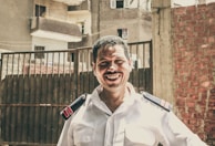 A man wearing a white uniform with epaulettes stands smiling in front of a gated area. The background consists of a mix of residential buildings and a brick wall, suggesting an urban setting. The lighting is bright, casting clear shadows on the man's face and uniform.