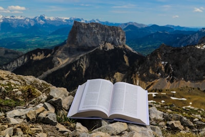 An open book rests on a rocky foreground, with a backdrop of expansive mountains and clear blue skies. The scene blends elements of nature with the act of reading, creating a serene and contemplative atmosphere.