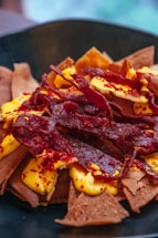 A close-up of a dish featuring crispy brown tortilla chips topped with a vibrant, creamy yellow cheese sauce and garnished with bright red, crunchy strips. The combination of colors and textures suggests a flavorful and visually appealing appetizer.