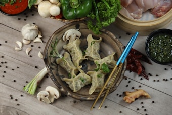 A rustic wooden surface is adorned with a bowl of cooked dumplings garnished with herbs. Surrounding the bowl are various ingredients including garlic, mushrooms, green peppers, spring onions, and a mix of spices. Nearby, there are vibrant red chili peppers, a dish of green sauce, and a steamer basket with raw meat. The setting suggests a preparation area for a flavorful meal.