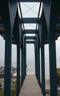 A wooden pathway leads through a structure of tall, dark blue columns with cross beams at the top. The pathway continues towards the open water, with rocks on the left and some bushes on the right.