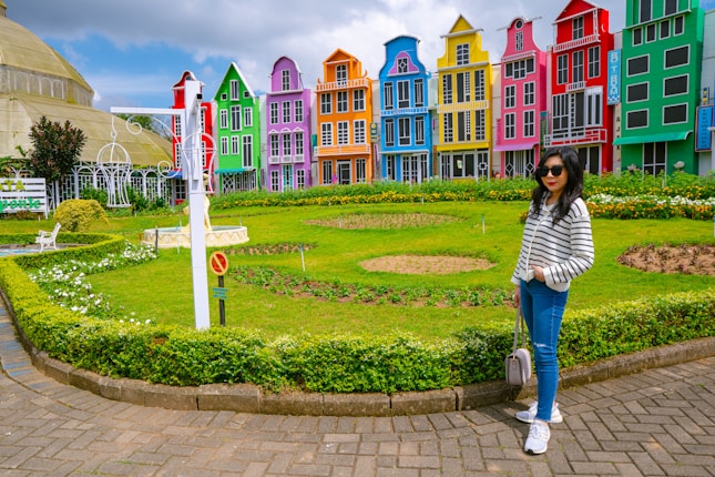 A colorful street scene with vibrant, multicolored buildings forming the backdrop. Each building is painted in a different hue, including red, green, blue, pink, yellow, and purple. A well-maintained green lawn and garden are in front, with a pathway of paving stones. A person wearing a striped top, jeans, and sunglasses stands casually, adding a human element to the bright and cheerful setting.