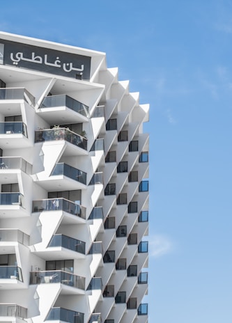 A modern building with a geometric facade featuring numerous balconies with glass railings. The structure has a repetitive pattern and the top of the building displays Arabic lettering. The sky is clear and blue.