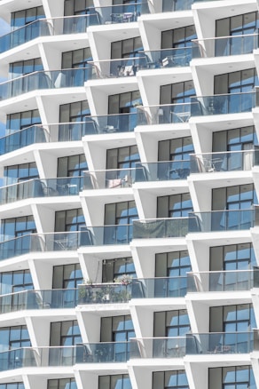 A modern architectural facade featuring a grid of geometric balconies with transparent glass railings. Each balcony has a similar design, though some include plants or furniture. The building is clad in white with large, rectangular windows allowing views into the apartments.