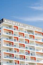 A modern apartment building features a series of stacked balconies with glass railings. The building facade incorporates a repeating pattern with white and orange color accents. The name 'BINGHATTI' is prominently displayed at the top in both English and Arabic.