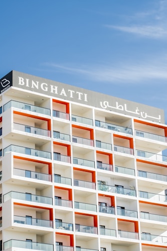 A modern apartment building features a series of stacked balconies with glass railings. The building facade incorporates a repeating pattern with white and orange color accents. The name 'BINGHATTI' is prominently displayed at the top in both English and Arabic.