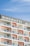 The image features a modern multi-story building with a prominent sign displaying 'Binghatti' at the top. The structure has a repetitive design with square balconies, each having glass railings and a distinctive orange back wall. The building stands against a clear blue sky.