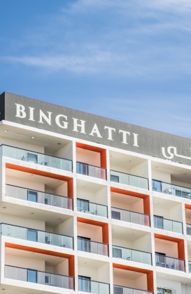The image features a modern multi-story building with a prominent sign displaying 'Binghatti' at the top. The structure has a repetitive design with square balconies, each having glass railings and a distinctive orange back wall. The building stands against a clear blue sky.