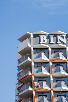 A modern high-rise building with geometric balconies and glass railings. The structure features a combination of white, grey, and orange colors with the top part displaying large block letters 'BIN'. The blue sky serves as the backdrop.
