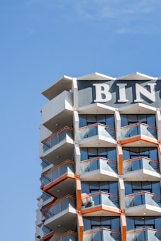 A modern high-rise building with geometric balconies and glass railings. The structure features a combination of white, grey, and orange colors with the top part displaying large block letters 'BIN'. The blue sky serves as the backdrop.